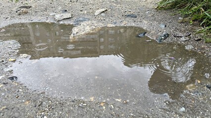 Puddle reflecting buildings and greenery, surrounded by gravel and stones, showcasing urban environment and natural elements with copy space for text overlay