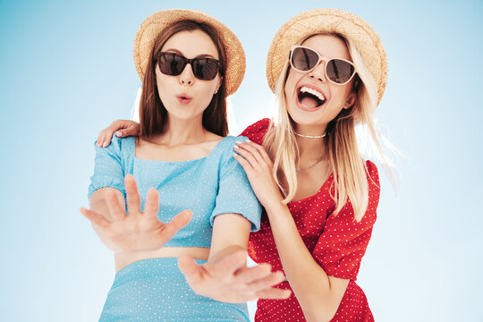 Two young beautiful smiling hipster woman in trendy summer same clothes. Carefree women in street in hats. Positive models at sunset. Cheerful and happy. Bottom view. They look at camera from above