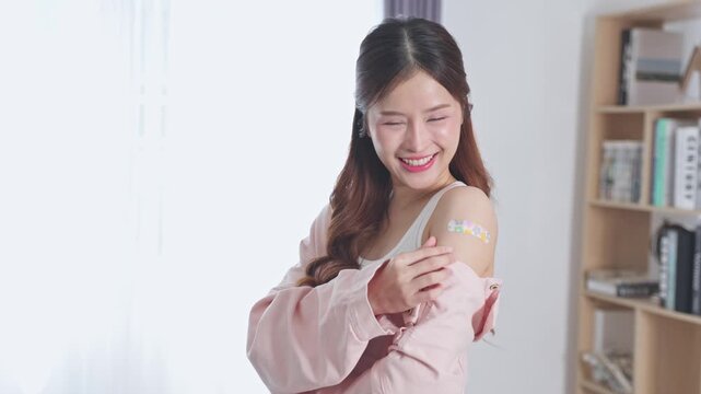 Asian Young woman with long hair smiling gently, colorful bandage on shoulder, wearing casual pink shirt indoors near bookshelf, showcasing health, vaccination, and positive emotion in bright room