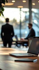 Modern office space shows two men working while another watches, laptop in foreground on desk