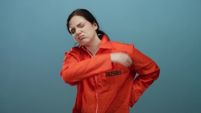 Woman wearing orange prison jumpsuit standing against blue background with a pained expression and hands on her back.