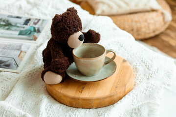 A teddy bear with a cup of coffee on a wooden tray, a cozy blanket, a wicker pillow and a magazine create an atmosphere of warmth and tranquility.
