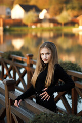A woman stands on a wooden bridge near a picturesque pond. She leans her back against the bridge railing, her pose is relaxed.
