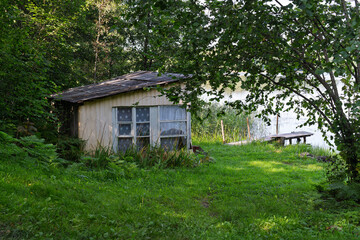 "Old wooden lakeside shed with curtained windows, surrounded by lush greenery and trees, with a small pier by calm water."