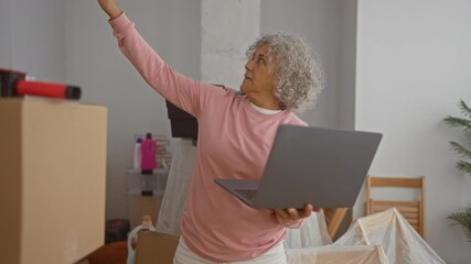 Grey-haired woman holding laptop during video call inside new home surrounded by moving boxes, conveying her age and domestic setting.