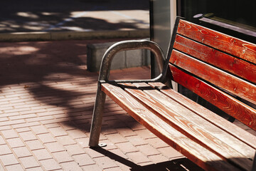 Empty wooden bench with metal armrests. Sunlight and shadow on red brick pavement. Outdoor seating in public space. Simple urban furniture design in city environment. Relaxation and waiting area.