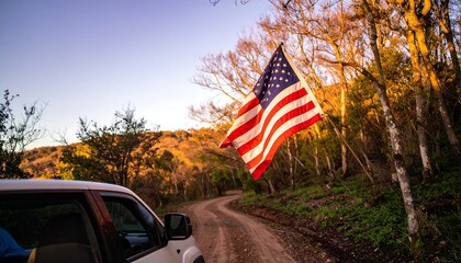A vibrant American flag waves gently over a dusty forest road, highlighting the golden hues of the late afternoon sunlight.