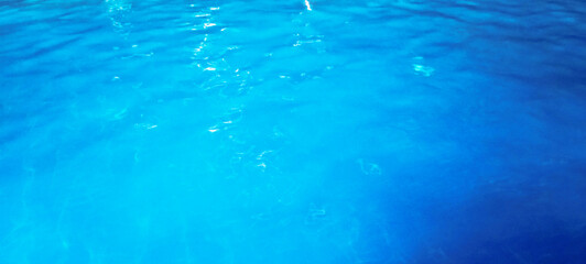 The crystal clear turquoise blue pool water surface gently ripples, reflecting sunlight and revealing the mosaic tiles beneath, creating a refreshing and inviting summer scene.