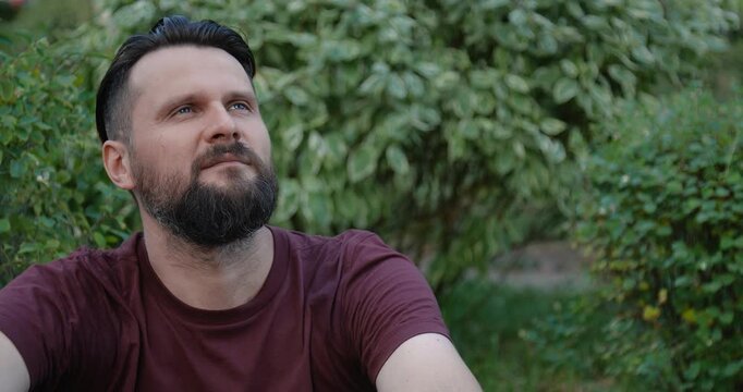 Close-up portrait of a bearded Caucasian man with an undercut hairstyle  straightening his hair looking to camera against a background of green leave