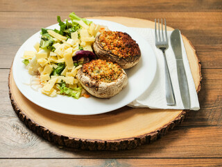 A plate of food with a fork and knife on a wooden table. The plate has a fresh salad with cheese and two fancy posh stuffed mushroom patties. Simple tasty meal.