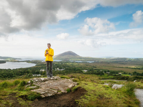Girl with smart phone standing on a rock, amazing nature scenery behind her with ocean, mountain and lush green fields of Connemara, Ireland. Travel and tourism. Model is slim in yellow jacket.