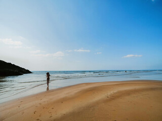A woman is walking on a beach into blue ocean water with a clear blue sky above her. The beach is sandy and the water is calm. Warm sunny summer vibe and mood. Surfers in the background.