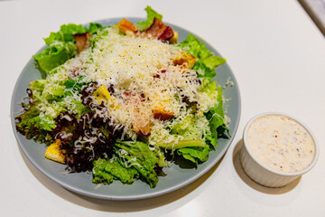 Close-up view of Caesar salad with shredded cheese, leafy greens, croutons, and bacon, served with creamy dressing in a small ramekin on the side
