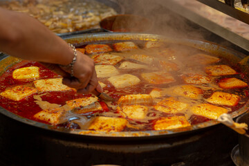 Street vendor stirring spicy Taiwanese braised tofu blocks in bubbling red chili broth at a night market food stall