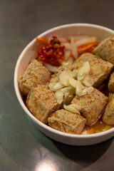 Crispy stinky tofu topped with sliced garlic and served with pickled vegetables and chili sauce in a paper bowl, photographed from above