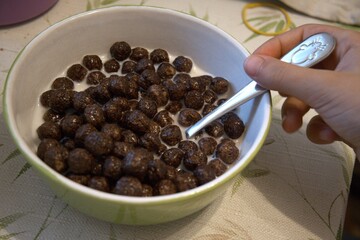 Crispy chocolate cereal balls served with milk in small bowl with hand, nutritious breakfast for children.