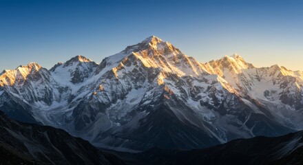 Majestic mountain range with snow-capped peaks illuminated by the golden light of sunrise or sunset against a clear blue sky.