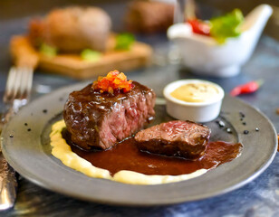 A plate of meat and potatoes with a sauce on the side. The meat is cut in half and the potatoes are cooked. The plate is on a wooden table