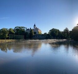 Fototapeta premium Morning light bathes the main building of Radun Castle in Opava, surrounded by serene lakes and lush nature