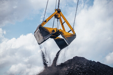 Heavy machinery is actively involved in unloading coal from a large bucket at an industrial location. The excavator crane, equipped with a dual clamshell bucket, hovers above a substantial pile of