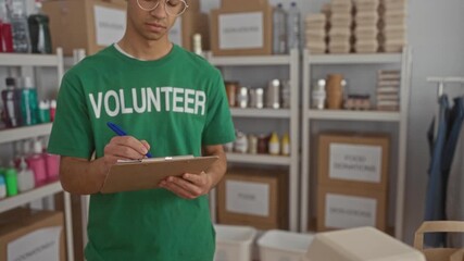 Young hispanic volunteer man wearing green shirt writes notes on clipboard in donation center room; compassion. - Powered by Adobe