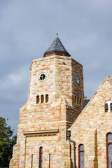 A beautiful historic stone church tower with a slate steeple and clock faces, captured against a moody cloudy sky. The warm-toned classic stonework, arched windows, and architectural details