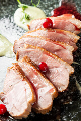Medium-rare duck breast slices with crispy skin, garnished with red berries and colorful sauces on a dark plate. Gourmet food presentation, close-up view