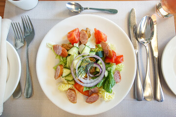  Salad with vegetables and sausage in a white plate, on a white napkin. Table setting in a restaurant. Top view.