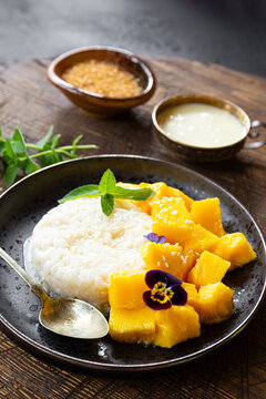Thai mango sticky rice with coconut milk, garnished with edible flowers and mint on wooden table vertical photo