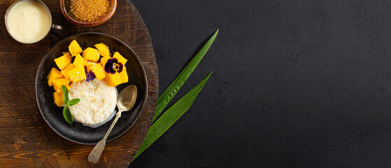 Thai mango sticky rice with coconut milk, garnished with edible flowers and mint on black background top view, web banner with copy space for text
