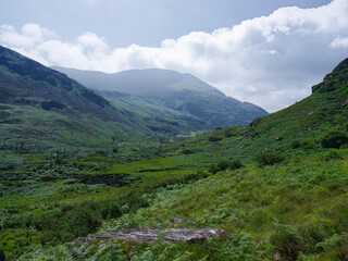 Fototapeta premium Rolling green hills fill the valley in Gap of Dunloe, Ireland. Mist hangs in the distance near the mountain tops under a partially cloudy sky, on a summer day.