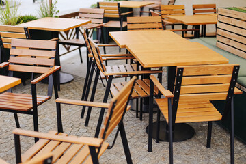 Wooden tables and chairs at outdoor cafe terrace. Empty restaurant furniture arranged on cobblestone pavement. Modern design with wooden seats and metal frames. Cozy atmosphere of urban dining area.