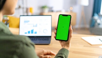 Modern workspace Person holds a green screen smartphone with laptop showing business analytics in the background, focusing on digital productivity and data management
