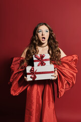 Fototapeta premium A shocked young woman in a gorgeous red dress holds holiday gifts amid festive cheer.