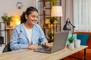 Young woman wearing headset, freelance worker, call center or support service operator helpline talking with client or colleague communication support. Girl using laptop at home office desk. Lifestyle