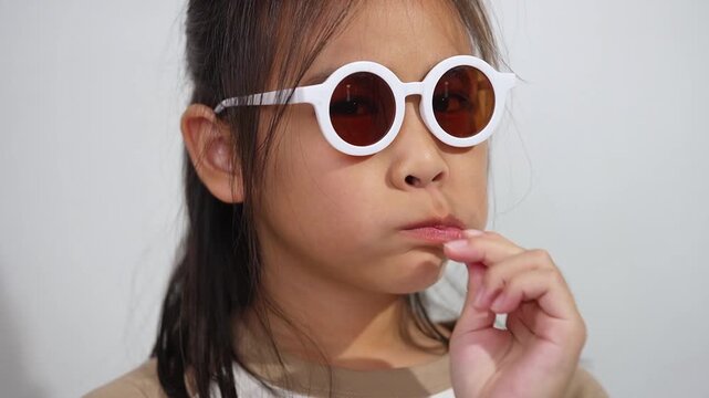 An Asian girl wearing round sunglasses while eating a lollipop, posing playfully with a fun and stylish expression against a white background.