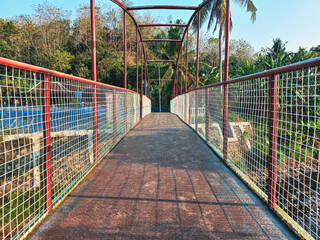 Obraz premium A striking red metal pedestrian bridge with a worn walkway and wire mesh fence leading through a lush green tropical forest on a bright sunny day