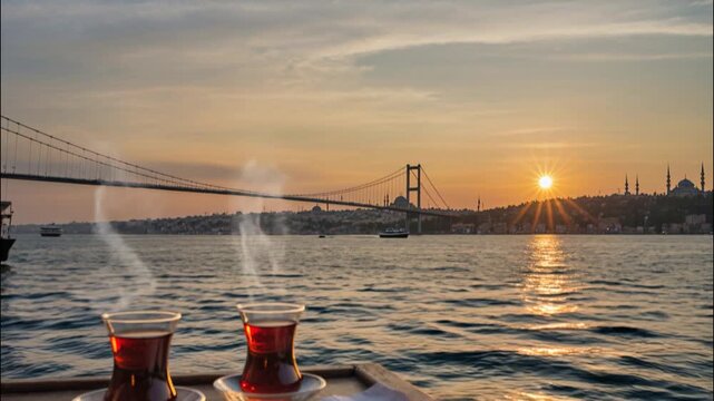 Turkish tea in traditional glasses with simit on a table by the Bosphorus at sunset, view of Istanbul skyline, Bosphorus Bridge, and historic mosques in the background.
