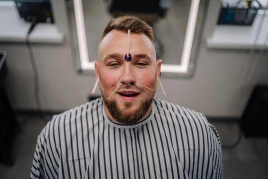 A smiling man sits in a barber chair with waxing sticks applied to his nostrils, upper lip, ears, and forehead during a grooming and hair removal session. - Powered by Adobe