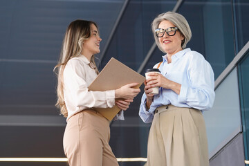 Two businesswomen are having a conversation outside their office building, holding coffee and documents