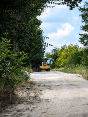 A bright yellow and black tractor parked on a sandy country road, framed by dense green foliage on both sides, under a clear summer sky with white fluffy clouds.