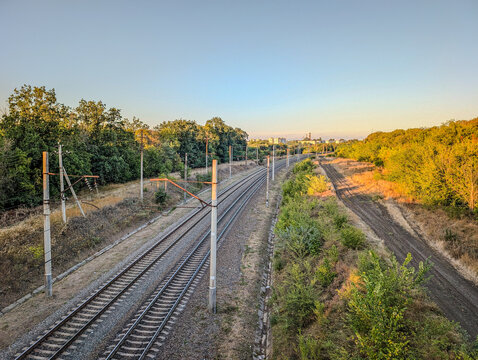 A wide, scenic view of two parallel railway tracks stretching into the distance through a rural landscape, with green forests and fields on both sides, and the warm, golden light of sunset