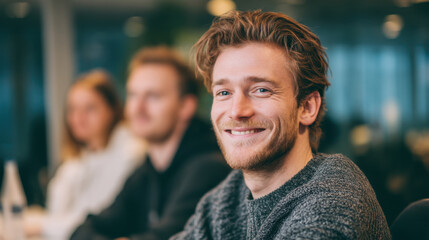 Fototapeta premium Young man with curly hair and beard smiles warmly while seated at table in modern office environment. atmosphere is professional and collaborative