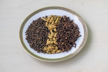 Black pepper, cardamom, and cloves on a white plate set on a light wooden surface, perfect for food, cooking, and spice-related projects.