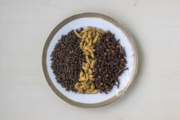 Assortment of black pepper, cardamom, and cloves on a white plate, resting on a wooden table. Spices used for flavoring culinary dishes. Top view.