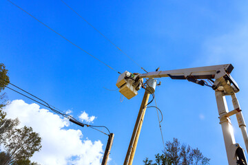 Workers use cherry picker to fix power lines under bright blue sky with fluffy clouds.