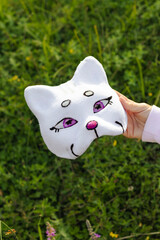 Close-up of white plush cat mask with purple eyes and pink nose held in hand over grass and...