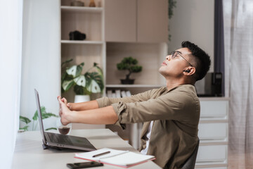 Asian man stretching arms upward while sitting at home office desk with laptop and coffee. Concept of healthy remote work routine, balancing productivity with physical wellness in modern lifestyle