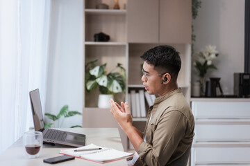 Fototapeta premium Asian man wearing glasses and earbuds gesturing hands while talking during online meeting at home office with laptop. Perfect for video calls, webinars, remote teamwork, and digital business concepts