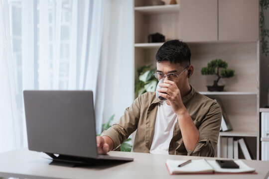 Asian man at home office desk using laptop while drinking coffee, smartphone and notebook beside him. Great for freelance, online business, and remote work productivity lifestyle themes